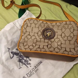 Beige axelväska från U.S. Polo Assn. - Säljer en beige axelväska från U.S. Polo Assn. Väskan har brunt mönster med logga och detaljer i mörkbrunt och gult. Justerbar axelrem i läderlook och dragkedja upptill. Perfekt storlek för det viktigaste.
