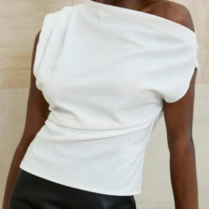 Vit offshoulder topp - Snygg vit topp med offshoulder-design och asymmetrisk halsringning. Toppens stilrena look och mjuka tyg gör den perfekt för dig som gillar minimalistisk stil. Passformen är normal och toppen är ärmlös, vilket ger en fräsch vibe.