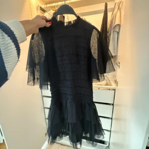 Svart kort klänning från Malina - Säljer en svart kort klänning från Malina med volangdetaljer och transparenta, korta ärmar. Klänningen har en cool struktur med flera lager och en feminin siluett. Perfekt för dig som gillar trendiga och unika plagg med lite edge.