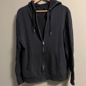 Svart hoodie med dragkedja Asos design - Svart hoodie från Asos design med hel dragkedja framtill, huva med snörning och två stora fickor. Tröjan är långärmad och har en loose passform, perfekt för chill dagar. Materialet är mjuk bomull som känns skönt mot huden.