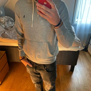 Grå hoodie från Polo Ralph Lauren - Säljer en grå hoodie från Polo Ralph Lauren med klassisk broderad logga på bröstet. Tröjan har huva med snörning, stor magficka och ribbade muddar. Perfekt för chill dagar och enkel att matcha med jeans eller joggers.
