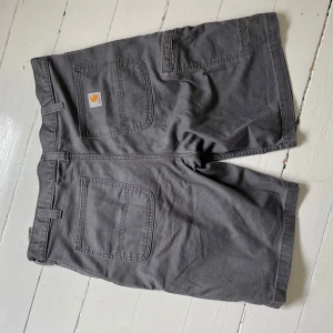 Grå Carhartt shorts relaxed fit - Snygga grå shorts från Carhartt i relaxed fit med raka ben och flera praktiska fickor. Klassisk design med bälteshällor och robust bomullsmaterial, perfekta för chill dagar eller när du vill ha en avslappnad stil.