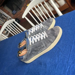 Blåa mocka sneakers från Saint Laurent - Snygga sneakers från Saint Laurent i blå mocka med vita snören och beige sula. Diskret logga på sidan och blå detaljer vid hälen och plösen. Perfekta för dig som gillar stilrena och exklusiva skor med en lyxig känsla.