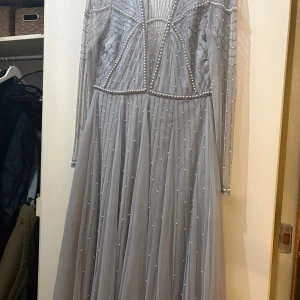 Ljusgrå pärlprydd långklänning ASOS - Säljer en magisk ljusgrå långklänning från ASOS DESIGN med transparent mesh, pärlprydda detaljer och glittriga broderier. Klänningen har lång ärm, hög hals och djup v-ringning framtill. Perfekt för dig som vill sticka ut med coola detaljer och elegant vibe.