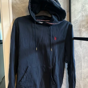 Marinblå hoodie från U.S. Polo Assn. - Säljer nu en marinblå hoodie från polo med röd logga broderad på bröstet. Tröjan har huva med snörning och en klassisk känguruficka framtill. Ränder i rött, vitt och blått på insidan av huvan ger en extra detalj.