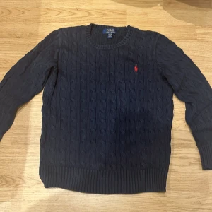 Mörkblå kabelstickad tröja Polo Ralph Lauren - Kabelstickad mörkblå tröja från Polo Ralph Lauren med klassisk röd broderad logga på bröstet. Tröjan har rund halsringning, långa ärmar och ribbade muddar. Perfekt för dig som gillar preppy och stilren look.