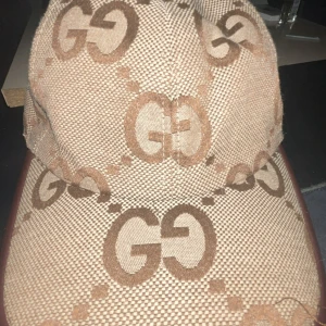 Beige keps från Gucci med GG-mönster - Säljer en beige keps från Gucci med det klassiska GG-logomönstret i bruna toner. Kepsen har böjd skärm och är tillverkad i bomullsmaterial med insida i ljus textil. Perfekt för dig som vill ha en lyxig och trendig accessoar med tydlig designerstil.