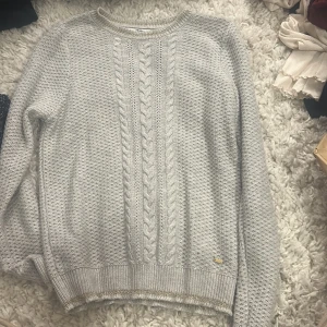 Ljusgrå stickad tröja Hampton Republic - Snygg ljusgrå stickad tröja från Hampton Republic i storlek S. Tröjan har ett klassiskt flätmönster framtill och ribbade muddar med en diskret glittrig detalj vid nederkanten. Perfekt för dig som gillar stilrena och mysiga plagg.