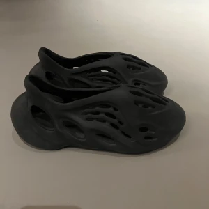 Svarta adidas Yeezy Foam Runner - Säljer ett par svarta adidas Yeezy Foam Runner med futuristisk design och många hål för ventilation. Skorna är tillverkade i syntetmaterial och har en rund tå samt platt sula. Perfekt för dig som gillar unik stil och komfort.