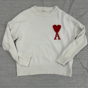 Vit stickad tröja från Ami  - Vit stickad tröja från Ami med ett stort rött hjärta och bokstaven A broderat på bröstet. Tröjan har rund hals, ribbade muddar och lång ärm. Perfekt för dig som gillar stilrena plagg med en twist. Materialet är mjukt och känns lyxigt. Dock väldigt använd