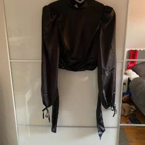 Svart satin bolerotopp med volang - Snygg svart bolerotopp i satin med långa puffärmar och knytband vid ärmsluten. Toppen har hög hals och öppen rygg med volangdetaljer längs kanterna.