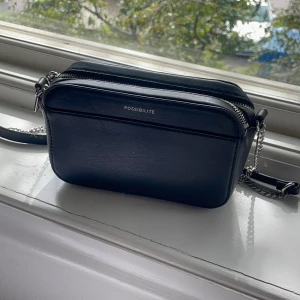 Svart axelväska  - Stilren svart axelväska från HM med silverfärgad kedjerem och dragkedja. Väskan är rektangulär och tillverkad i slätt skinnliknande material. Perfekt storlek för mobil, plånbok och andra essentials. Enkel och clean design med diskret logga framtill. Skriv om du undrar något 💕