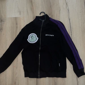  Moncler x Palm Angels sweater - Svart track jacket med dragkedja från Moncler x Palm Angels. Jackan har lila ränder längs ärmarna, broderad Palm Angels-logga på bröstet och stor Moncler-patch. Sportig vibe och ribbade muddar. Perfekt för dig som gillar streetwear och snygga detaljer.