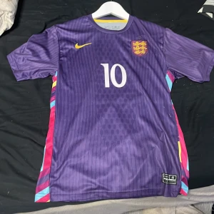 England Bellingham #10 lila fotbollströja Nike - Snygg England fotbollströja från Nike i lila med gula och färgglada detaljer på sidorna. Nummer 10 och Bellingham på ryggen. Tillverkad i Dri-FIT-material som andas. Kortärmad modell med klubbmärke och Nike-logga på bröstet.