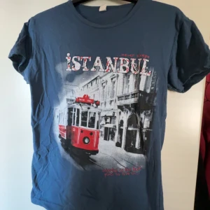 Blå Istanbul t-shirt  - Snygg blå t-shirt med coolt Istanbul-tryck framtill och motiv av en röd spårvagn. Klassisk rund hals och korta ärmar. Perfekt för dig som gillar unika tryck och streetstyle. Materialet känns som bomull och t-shirten har en avslappnad passform.