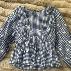 Blåvit rutig blus från Zara - Supersöt blus från Zara i blåvitt rutigt mönster med broderade vita blommor. Blusen har v-ringning, lång ärm och volang nedtill som ger en extra touch. Tillverkad i lätt bomull, perfekt för vår och sommar. 