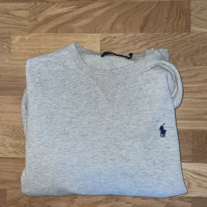 Grå sweatshirt från Polo Ralph Lauren - Snygg grå sweatshirt från Polo Ralph Lauren med klassisk blå broderad logga på bröstet. Tröjan har rund hals och är tillverkad i mjuk bomull, perfekt för chill dagar. Enkel och stilren design som funkar till jeans eller joggers.