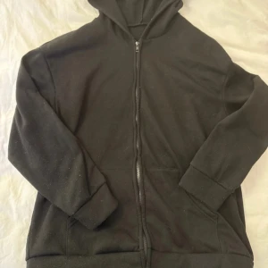 Svart hoodie med dragkedja - Svart hoodie med huva och hel dragkedja framtill. Tröjan har långa ärmar och två fickor på magen. Perfekt för chill dagar eller när du vill ha något bekvämt och enkelt. Materialet känns mjukt och är troligtvis bomull eller polyester.