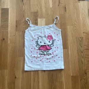 Hello Kitty vit linnetopp med hjärtan - Supersöt vit linnetopp med smala axelband och ett stort Hello Kitty-tryck framtill. Toppen har rosa hjärtan runt motivet och en rosa blomma i håret på Hello Kitty. Perfekt för dig som älskar gulliga detaljer och vill ha en lekfull stil.