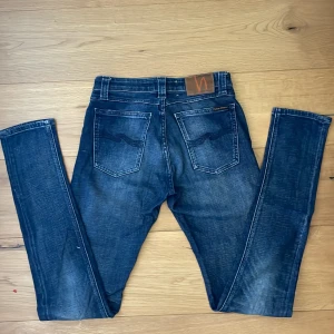 Nudie Jeans Skinny Blå 26/34 - Säljer ett par snygga skinny jeans från Nudie Jeans i mörkblå tvätt med slitningar. Klassisk femficksmodell med orange sömmar och Nudie-logga på bakfickan. Jeansen är i stretchig bomull och har smal passform hela vägen ner.