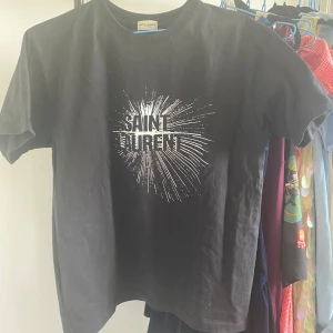 Svart Saint Laurent t-shirt - Svart t-shirt från Saint Laurent med coolt vitt tryck framtill som sprakar ut från loggan. Klassisk rund hals och korta ärmar. Skön passform och tillverkad i mjuk bomull. Perfekt för dig som gillar streetstyle och vill ha en ikonisk look. Skriv vid funderingar!