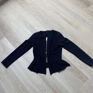 Svart kavajjacka från Gina Tricot - Svart kavajjacka från Gina Tricot med dragkedja framtill och volangdetalj nedtill. Jackan har lång ärm och figurnära passform, perfekt för dig som gillar en stilren look med en twist. Materialet är mjukt och stretchigt för extra komfort.