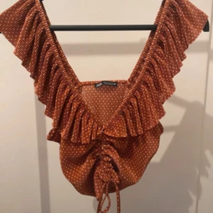 Prickig volangblus från Zara - Supersöt brun blus från Zara med vita prickar, djup v-ringning och breda volanger längs axlarna. Toppen har snörning framtill som ger en snygg rynkad effekt och croppad passform. Perfekt för dig som gillar unika detaljer och trendig stil.