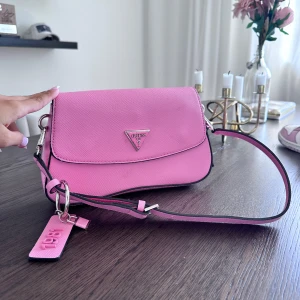 Rosa axelväska från Guess - Snygg rosa axelväska från Guess med silverfärgad logga framtill och matchande axelrem. Väskan har en stilren design och en tag med '1981' i rosa. Perfekt för dig som vill ha en poppig accessoar till din outfit.