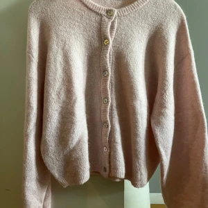 Ljusrosa kofta från Gina Tricot - Supermjuk och fluffig ljusrosa kofta från Gina Tricot. Koftan har långa ärmar, ribbade muddar och knäppning med fina knappar framtill. Perfekt för lager-på-lager och ger en mysig vibe till din outfit. Använd endast ett fåtal gånger och är som ny! 