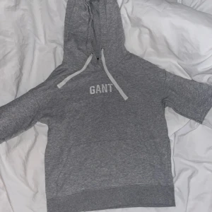 Grå hoodie från GANT med orange detalj - Snygg grå hoodie från GANT med vit logga på bröstet. Tröjan har huva med snörning och coola vita och orangea detaljer på ärmarna. Perfekt för en chill och sportig look. Mjuk och skön bomullskänsla, passar till allt.