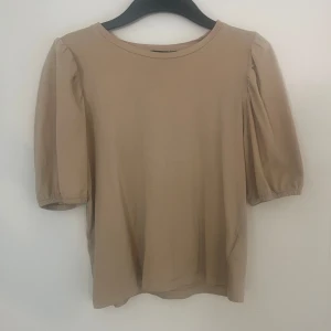 Beige topp med puffärm - En stilren beige topp med puffiga korta ärmar och rund halsringning. Tillverkad i mjuk bomull, perfekt för en enkel men trendig look. Passar till både jeans och kjol för en avslappnad vibe.