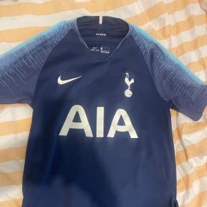Tottenham Hotspur blå matchtröja Nike - Snygg Tottenham Hotspur fotbollströja från Nike i mörkblått med ljusblå detaljer på ärmarna. Tröjan har klubbmärke och Nike-logga på bröstet samt AIA som sponsortryck. Tillverkad i lätt och ventilerande material, perfekt för träning eller match. INGEN SPELARE PÅ RYGGEN