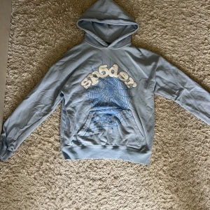 Ljusblå hoodie från Sp5der med strass - Ljusblå hoodie från Sp5der med stor spindelnätsgrafik och broderad logga framtill. Texten är dekorerad med strassdetaljer som ger extra bling. Klassisk känguruficka och huva. Tillverkad i 100% bomull för skön känsla. Pris kan diskuteras 