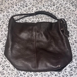 Brun axelväska i äkta skinn med fjärilsdetalj - Säljer en stor brun axelväska i äkta skinn med grov struktur. Väskan har en bred axelrem, dekorativ fjärilsdetalj i läder och praktisk dragkedja. Insidan är rymlig och har kedjedetaljer. Perfekt för dig som vill ha plats för allt du behöver.
