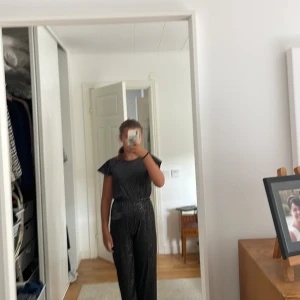 Svart glittrig byxdress från Lindex - Snygg svart byxdress från Lindex med glittriga ränder och vida ben. Byxdressen har korta volangärmar och markerad midja. Perfekt för dig som vill sticka ut med en cool och trendig look.