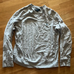 Grå sweatshirt från Polo Ralph Lauren - Säljer en stilren grå sweatshirt från Polo Ralph Lauren med klassisk rund hals och broderad grön logga på bröstet. Tröjan är i mjuk bomullsblandning med ribbade muddar och passar perfekt till jeans eller joggers.