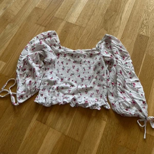 Vit blommig topp - Söt vit (lite croppad) topp med små rosa blommor från New Yorker. Överkroppen är sydd i smock och ärmarna är halvlånga med en rosett detalj nedtill. Den går att ha både on och off shoulder och har en liten volangkant nedtill. Toppen är i storlek xs men funkade för mig även när jag normalt hade s. Säljer då den nu blivit för liten.  Jag säljer även en topp i samma modell men annan färg om det är av intresse. Hör gärna av dig om du har frågor! 😊