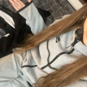 Grå Nike hoodie med dragkedja - Säljer en grå hoodie från Nike med svart dragkedja och snörning i huvan. Tröjan har en sportig vibe och är perfekt för chill dagar eller när du vill vara bekväm. Klassisk Nike-logga på bröstet och mjukt material.
