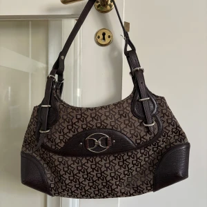 Brun axelväska från DKNY med monogram - Snygg axelväska från DKNY i brunt med klassiskt monogrammönster. Väskan har detaljer i mörkbrunt skinn, justerbara axelremmar med spännen och ett silverfärgat DKNY-emblem framtill. Invändigt finns ett fack med dragkedja och svart textilfoder.