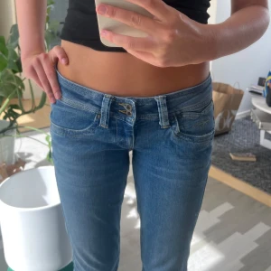 Blå lågmidjade bootcut jeans - Säljer ett par blå jeans med låg midja och bootcut-ben. Jeansen har klassisk femficksdesign, snygga slitningar och dubbla knappar i midjan. Perfekta för dig som gillar en avslappnad och trendig look.
