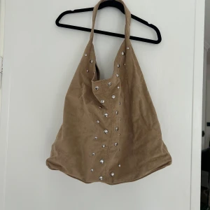 Beige axelväska med nitar - Säljer en stor och mjuk axelväska i beige mockaimitation med coola silvriga nitar framtill. Väskan har en bred axelrem och rymlig insida, perfekt för dig som vill ha plats för allt. Enkel men unik design som lyfter vilken outfit som helst. Pris kan diskuteras vid snabb affär