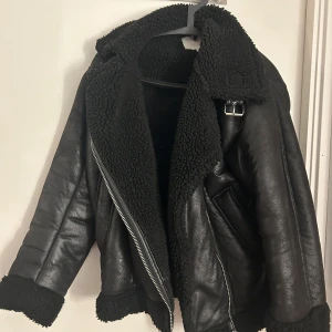 Svart teddyjacka med skinnlook - Snygg svart teddyjacka med skinnimitation och fluffigt foder. Jackan har bred krage, dragkedja i silver och coola detaljer som spänne vid halsen. Perfekt för dig som vill ha en trendig och varm jacka med edgy vibe.