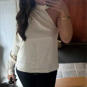 Vit oneshoulder blus med hög krage - Säljer en stilren vit blus med oneshoulder-design och hög krage. Toppen har en lång ärm med bred manschett och är tillverkad i ett glansigt material som ger en lyxig känsla. Perfekt för dig som vill sticka ut med en unik och trendig look.