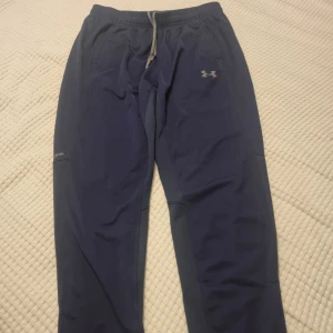 Blå träningsbyxor från Under Armour - Mörkblå mjukisbyxor från Under Armour med elastisk midja och grå snörning. Byxorna har raka ben, sidofickor och logga på låret. Se gärna mina andra annonser och skapa bundles. Se bilder för mått.