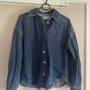 Jeansskjorta med pärlor från Zara XS - Snygg jeansskjorta från Zara i mörkblå denim med silverfärgade pärlor framtill. Skjortan har klassisk krage, långa ärmar och knappar framtill. Perfekt för dig som vill sticka ut lite extra med detaljer på en klassisk jeanslook.