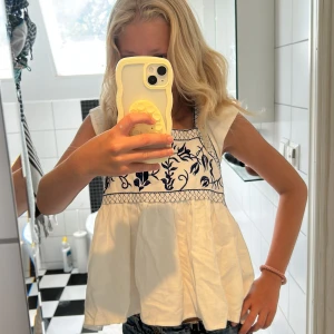 Vit blus med blå broderi och knyt - Supersöt vit blus med blå broderade detaljer och volangärmar. Toppen har en luftig passform, fyrkantig ringning och knyt i ryggen. Perfekt för varma dagar.