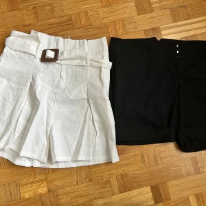 2-pack shorts: vita linne & svarta H&M - Säljer två par shorts! Ett par vita, vida shorts i linne med hög midja, bälte och snyggt spänne. Det andra paret är svarta, klassiska shorts från H&M med uppvikta ben och midwaist passform. Perfekta för sommaren och lätta att matcha.