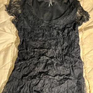 Svart spetstopp från Kappahl - Supersnygg svart topp från Kappahl med transparenta spetsdetaljer och lätt glittrig yta. Klänningen har korta ärmar och en rund halsringning. Perfekt för dig som gillar en edgy och feminin stil.