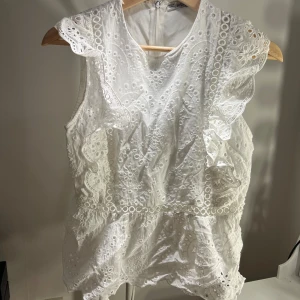 Vit broderad blus med volang - Superfin vit blus i bomull med broderade detaljer och volanger längs axlarna. Ärmlös modell med rund halsringning och vackert hålmönster framtill. Perfekt för en fräsch och somrig look!