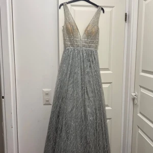 Kimalteleva hopeanvärinen pitkä mekko, jossa syvä v-pääntie - Taianomainen pitkä mekko hopeanvärisenä, jossa on kimalteleva kuvio ja syvä v-aukko. Mekossa on leveät olkaimet ja helmaosa. Kimalteleva kangas antaa todella ylellisen tunnelman. Täydellinen niille, jotka haluavat erottua joukosta ja loistaa tanssiaisissa tai juhlissa!
